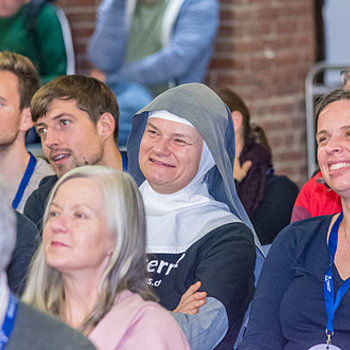 Blick ins Publikum, Menschen sitzen, im Fokus eine Nonne/Diakonisse, die freundlich lacht