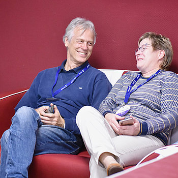 Mann mit grauen Haaren, schlank sitzt lässig mit Frau im Freizeitlook auf einem Sofa, beide lachen.