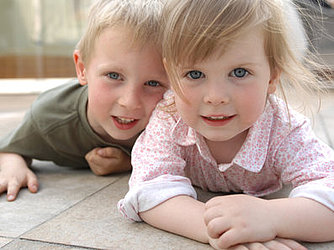 Zwei Kleinkinder krabbeln auf dem Boden und schauen in die Kamera.