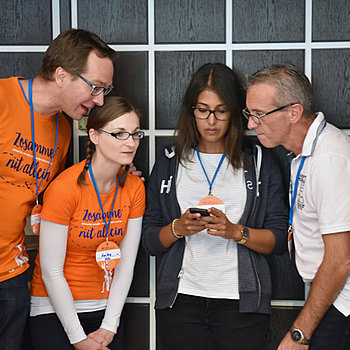 Zwei Frauen und zwei Männer schauen gemeinsam auf ein Handy, zwei haben Team-T-Shirts in Orange an.