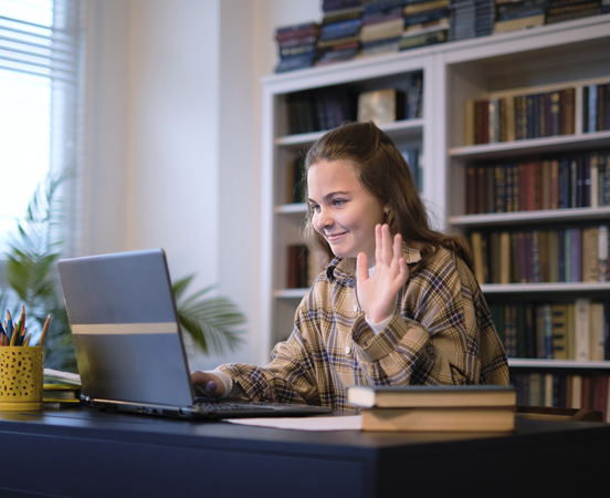 Junge Frau sitzt am Schreibtisch vor einem Laptop, winkt in die PC-Kamera, Bücherregal im Hintergrund